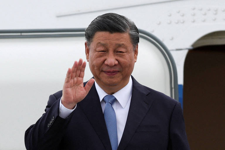 Retrato de Xi Jinping, um homem idoso, amarelo, com cabelos lisos, curtos e pretos, com algumas mechas brancas; ele veste terno preto, camisa branca e gravata azul; ele acena