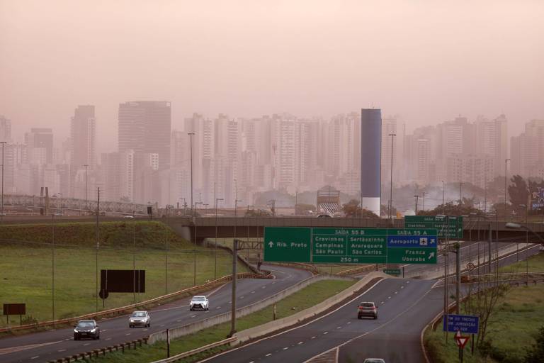 foto mostra via expressa larga, de duas mãos, placas de trânsito e alguns carros. ao fundo, é possível ver prédios através de nuvem espessa de poeira 