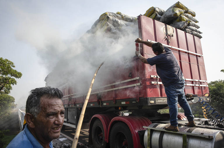 Caminhão com fumaça em sacos de carvão; ao lado há um homem com um bambu na mão e outro homem mexe na carga