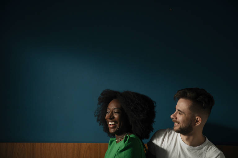 Foto de um casal de uma mulher preta e um homem branco, ela veste uma blusa verde e ele uma blusa branca, a parede ao fundo tem baixa iluminação, na cor azul.