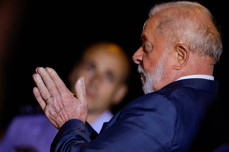 Lula, um homem branco de barba e cabelos da mesma cor, está de perfil, vestido com terno azul, as palmas das mãos juntas diante do corpo. Ao fundo, um homem careca de terno azul aparece desfocado