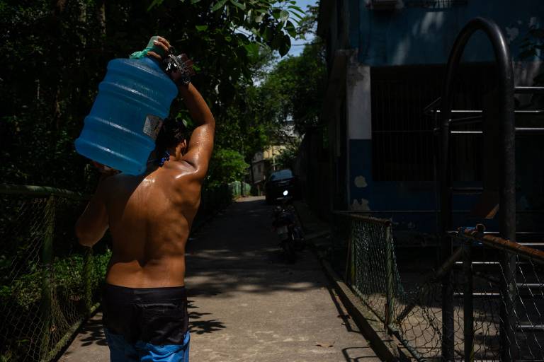 homem sem camisa, usando bermuda, carrega galão de água nas costas em viela