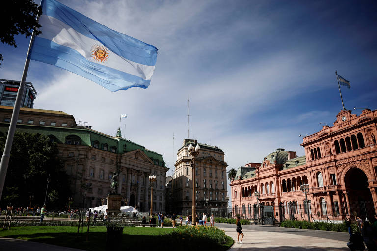 A imagem mostra a bandeira da Argentina tremulando em um mastro, com edifícios históricos ao fundo. À esquerda, há um edifício verde e, à direita, um prédio de arquitetura clássica em tons de rosa. O céu está parcialmente nublado, com algumas nuvens visíveis.