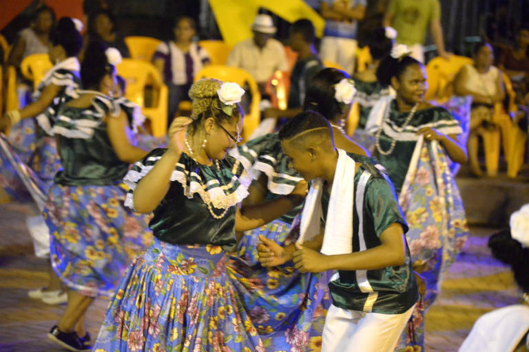 mulheres de saias coloridas dançam com homens ao fundo em praça à noite