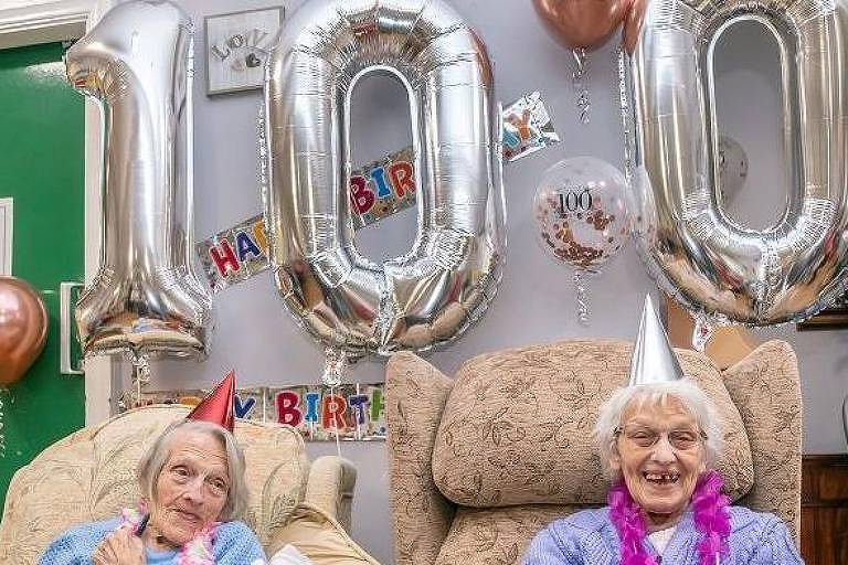 Duas senhoras sentadas sorrindo em frente a balões em formato de 100
