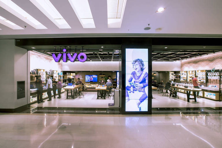 Fachada de loja da Vivo (o logo da marca, em roxo, está no alto da entrada) em interior de shopping. Há uma grande tela vertical na fachada, com a imagem de uma pessoa