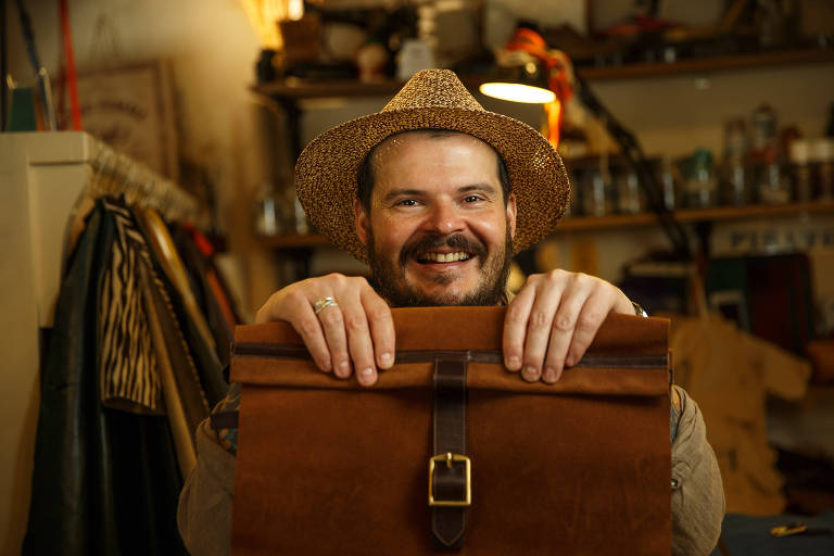 Homem branco de chapéu e sorridente segura com as ambas as mãos uma bolsa de couro marrom