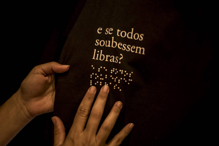 A imagem mostra uma mão fazendo a leitura de braile em uma camisa onde há a frase 'e se todos soubessem libras?'; a camisa é da cor preta