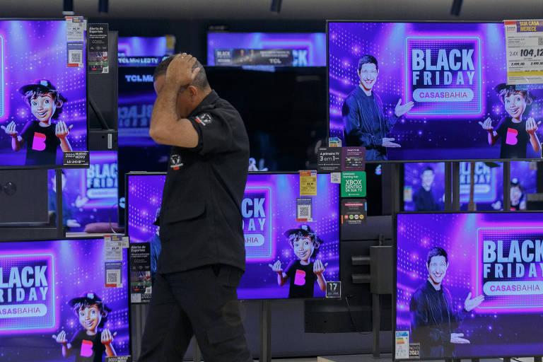 Homem branco, cerca de um e oitenta metros de altura, vestindo camisa de manga curta e calça preta, pasasando com a mão na nuca em frente de diversos televisores de diferentes polegadas e que mostram imagens roxas contendo a frase Black Friday casas bahia