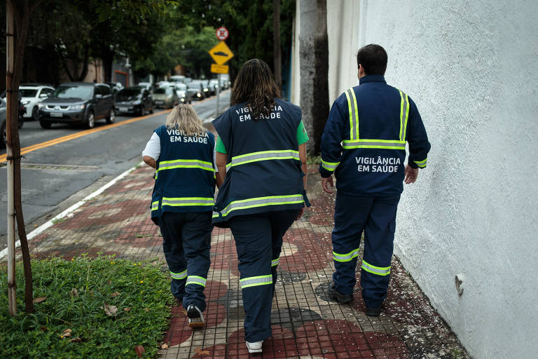 Três profissionais de saúde caminham pela calçada, usando uniformes azuis com faixas refletivas. As costas de dois deles são visíveis, e um deles é um homem. O ambiente é urbano, com carros estacionados e uma rua ao fundo. O texto 'VIGILÂNCIA EM SAÚDE' está visível nas costas dos uniformes.