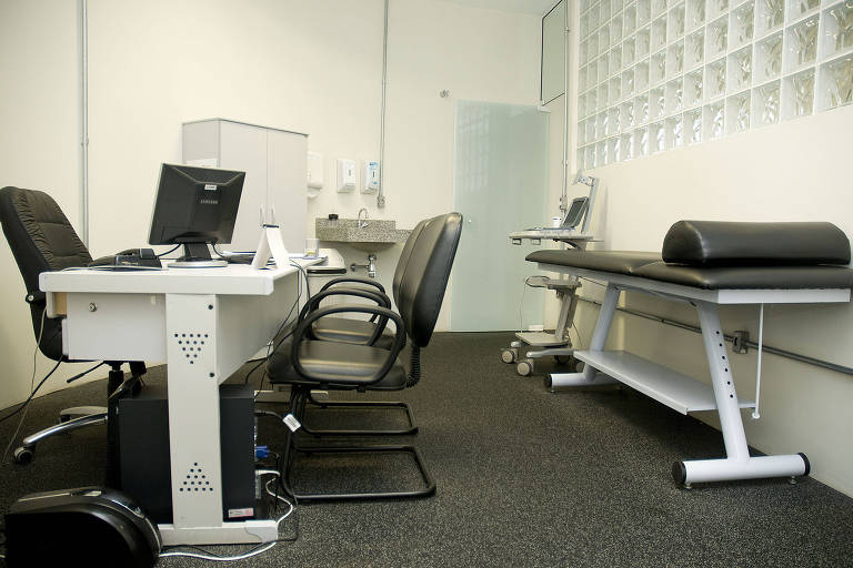 Sala de consultório médico com maca branca com acolchoado preto encostada à parede e mesa branca a frente, com tela de computador em cima da mesa. Há 3 cadeiras, dispostas duas frente a uma, todas na cor preta.
