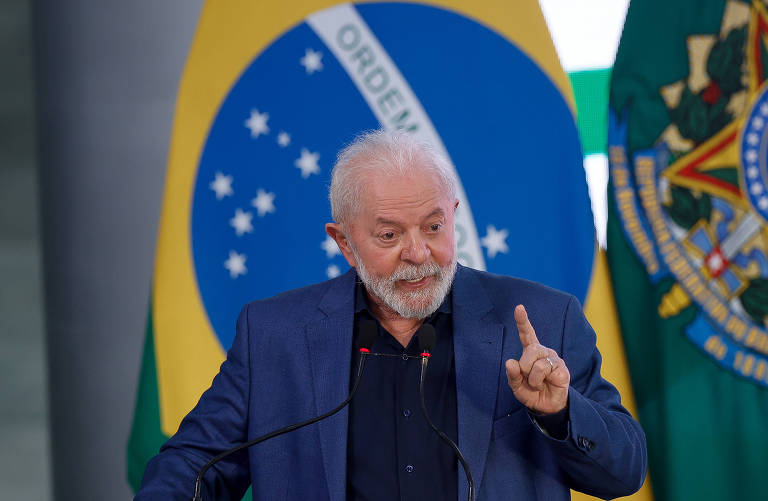 O presidente Lula vestido de terno e camisa azul, discursando na frente de um microfone preto. Ao fundo, temos a bandeira nacional do Brasil e bandeira presidencial do Brasil.