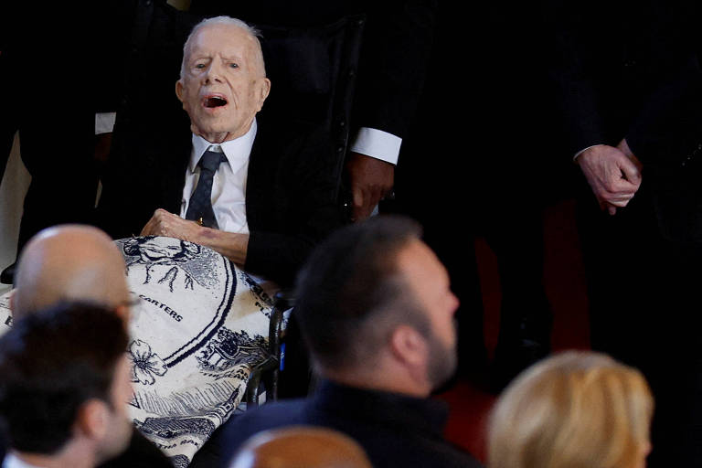 O ex-presidente dos EUA Jimmy Carter, que está sob cuidados paliativos, durante cerimônia em homenagem a sua esposa, Rosalynn, morta no último dia 19; em seu colo, uma manta com o rosto dela bordado