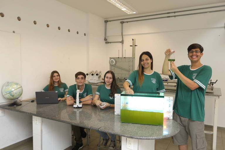 Imagem mostra alunos mexendo com equipamentos da pesquisa. Eles usam um uniforme verde escuro que sinaliza a equipe. Sobre a mesa, é possível ver um aquário com água verde, onde as algas estão armazenadas. Eles também tem um microscópio e um laptop sobre a mesa.