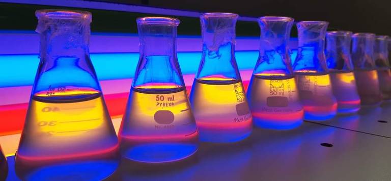 imagem mostra vidros de laboratório transparentes irradiados por uma luz amarela, rosa, vermelha e azul