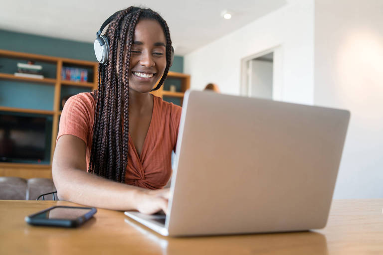 .Fotografia de uma jovem negra com tranças trabalhando e sorrindo em frente a um computador