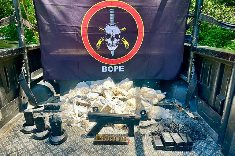 Na imagem, uma submetralhadora desmontada. Ao fundo, uma bandeira do Bope, com a ilustração de uma caveira e duas pistolas atrás