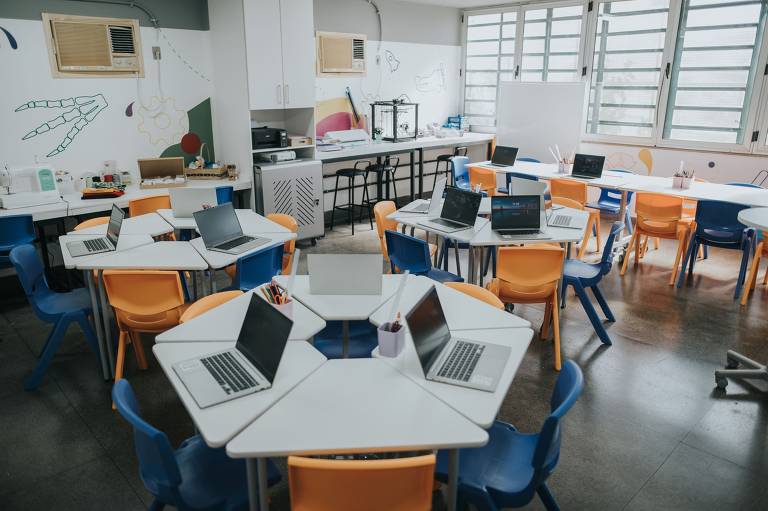 Sala de aula vazia é mostrada com diversas mesas e nelas diversos notebooks