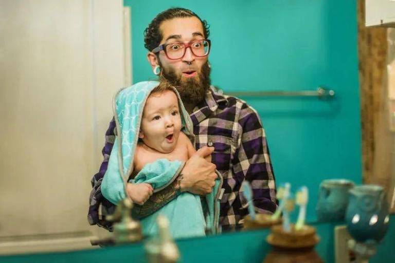 Homem com bebê no colo; eles olham no espelho e fazem caretas