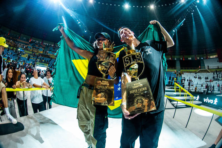 Os dois estão lado a lado, cada um com seu troféu; ambos sorriem e seguram juntos uma bandeira do Brasil nas costas