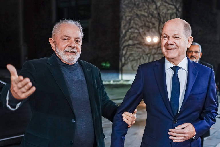 Homem branco idoso de barba usando blazer escuro gesticula, sorrindo, ao lado de homem branco idoso careca