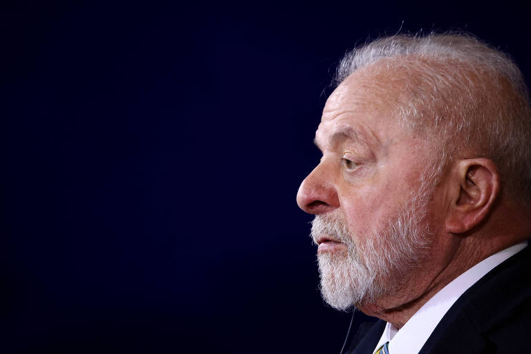 Lula, um homem branco de cabelo e barba careca, aparece de perfil, da região do ombro para cima. Ele usa terno preto e camisa branca