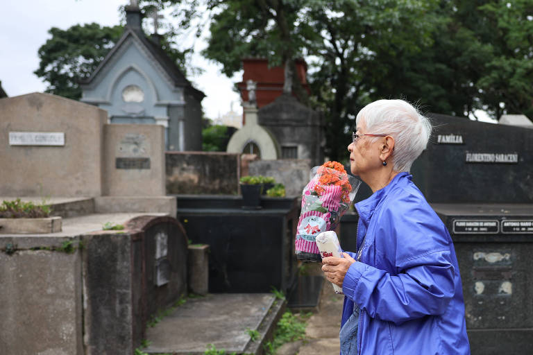 mulher de cabelos brancos segura um vaso de flor próximo de um túmulo
