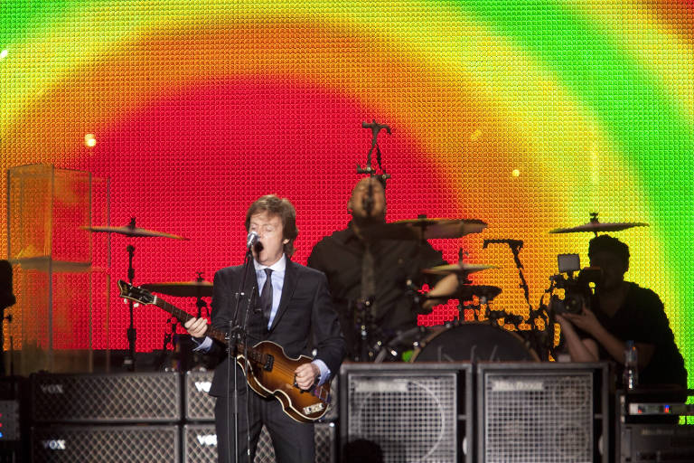 Um homem de terno canta ao microfone tocando uma guitarra, ao fundo, uma imagem que mescla cores como vermelho, amarelo, verde e vermelho