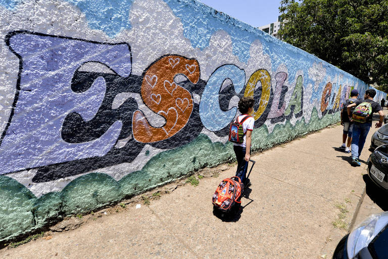 Criança caminhando com uma mochila de rodinha em frente a um muro colorido com a palavra "Escola"