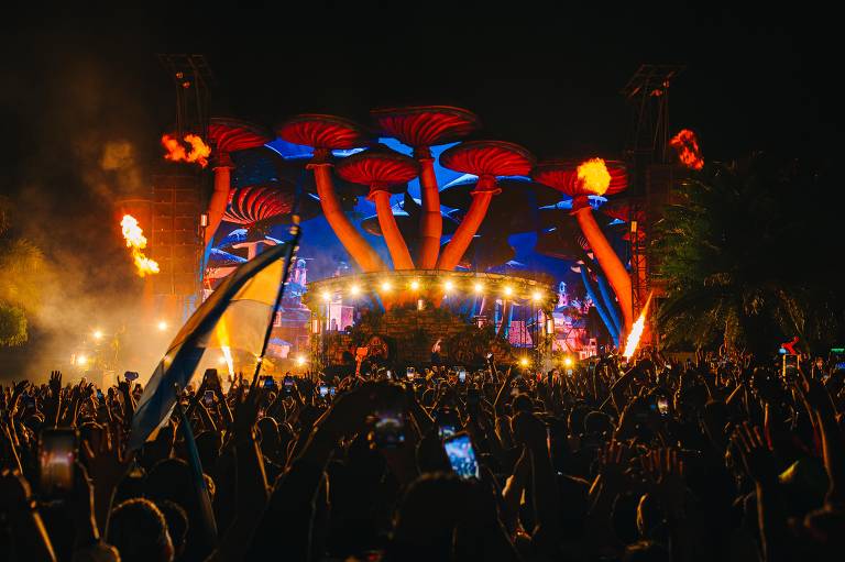 A imagem mostra um grande público em um festival de música à noite, com muitas pessoas levantando as mãos. Ao fundo, um palco iluminado com estruturas em forma de cogumelos vermelhos e azuis, além de chamas saindo do palco. O ambiente é vibrante e festivo, com luzes coloridas e uma atmosfera animada.