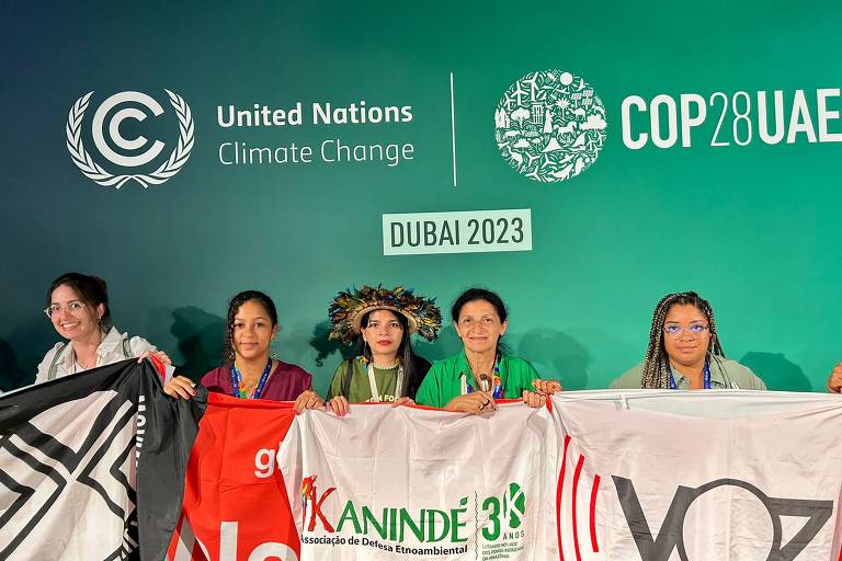 cinco mulheres indígenas posam atrás de bandeira na cop28