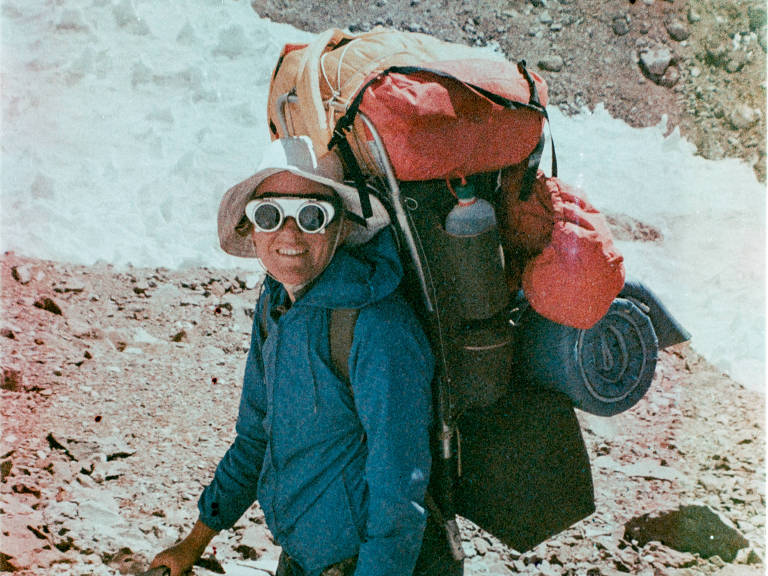 Alpinista com casaco azul, várias malas nas costas e óculos de sol