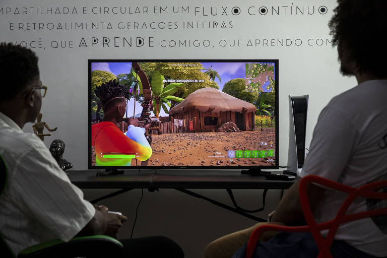 Duas pessoas de pele negra, uma mais clara e outra mais escura, jogando videogame juntas em uma sala com uma TV e um console de videogame