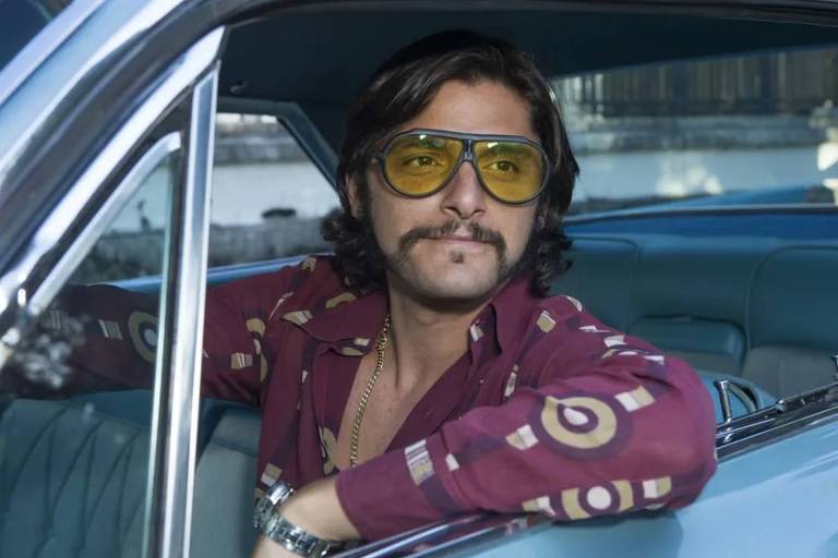 Em foto colorida, homem de camisa vinho e óculos escuros aparece sentado no banco da frente de um automóvel