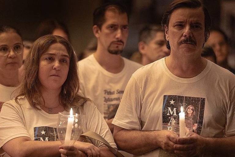Várias pessoas estão em vigília, incluindo Débora Lamm como Suzana e Thelmo Fernandes como Pedro, que seguram velas acesas e usam camisetas com foto da filha morta, em cena da série "Todo dia a mesma noite"