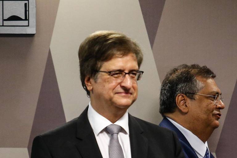 Paulo Gonet, de frente na altura dos ombros, e Flávio Dino, de perfil, no Senado Federal. Os dois vestem terno e usam óculos