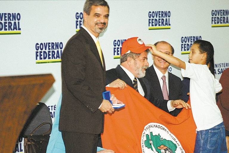 Uma criança coloca um boné do MST no presidente Lula. Dois homens seguram a bandeira do MST em frente ao corpo de Lula