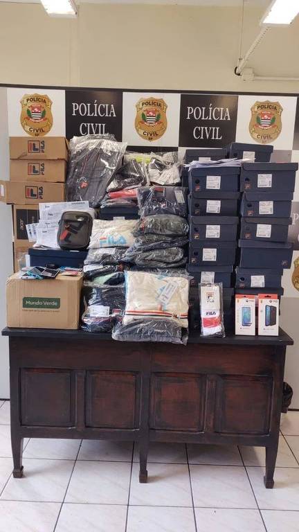 Mesa tem roupas, celulares e computadores empilhados à frente de banner da polícia civil