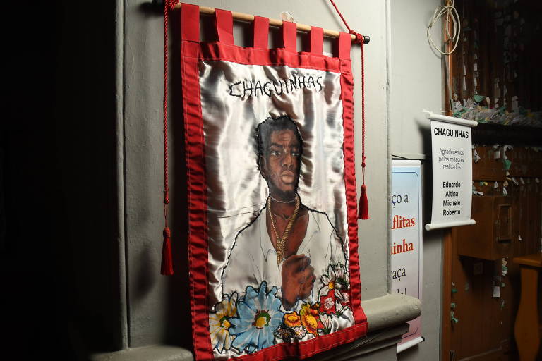foto colorida mostra estandarte com borda vermelha e a imagem de um homem negro com uma corda de forca ao redor do pescoço. O nome "Chaguinhas" e flores também enfeitam a bandeira