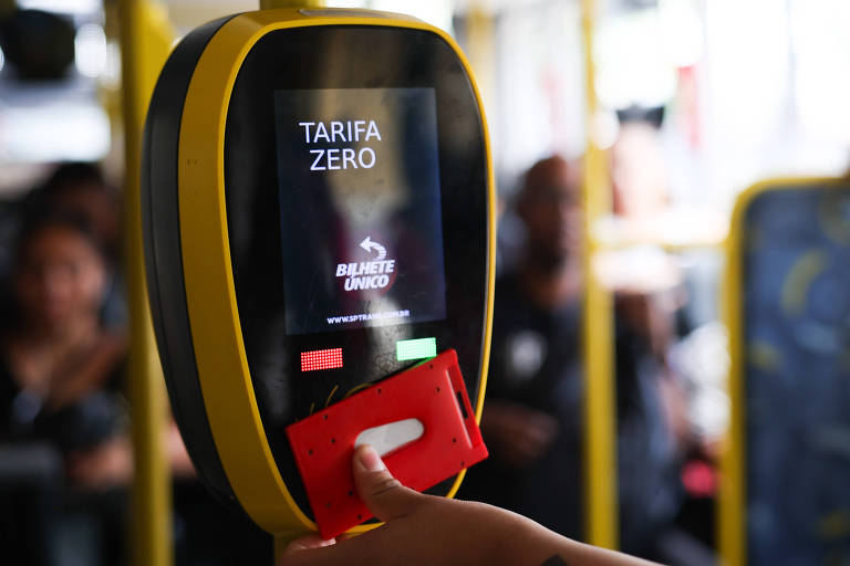 A imagem mostra um validador de passagem em um ônibus， com uma tela que exibe a mensagem 039;TARIFA ZERO039;. Uma mão está segurando um cartão vermelho próximo ao validador. Ao fundo， é possível ver outras pessoas dentro do ônibus.