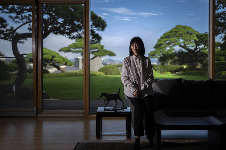 Koo Koo Yeon-kyung é uma mulher amarelha, de cabelos lisos e pretos, camisa branca, calça social e botas pretas.