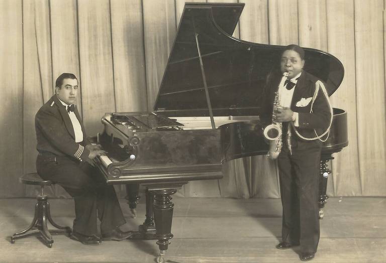 Em foto preto e branco, o saxofonista Euclydes de Araujo Senna aparece segurando um saxofone ao lado de um pianista e seu instrumento 