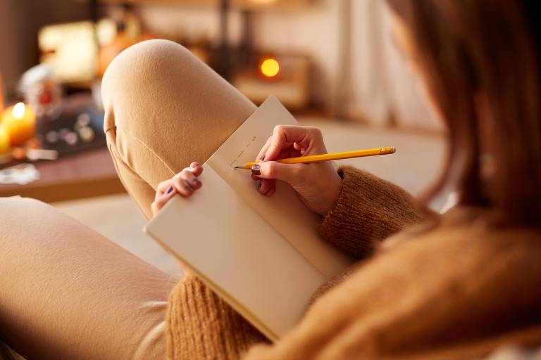 Fotografia colorida mostra uma mulher branca de cabelos ruivos sentada em um sofá escrevendo em uma agenda