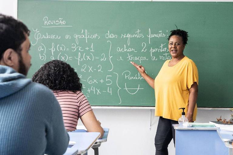 Foto mostra professora negra de pé dando aula para uma classe de alunos jovens e diversos de costas