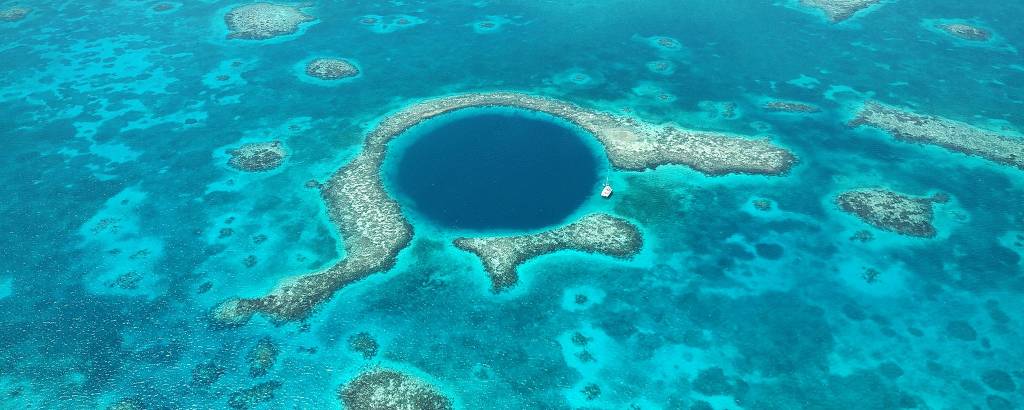 Vista aérea do recife, com um grande buraco circular ao meio, de um azul escuro. Junto à borda do buraco há um navio branco estacionado