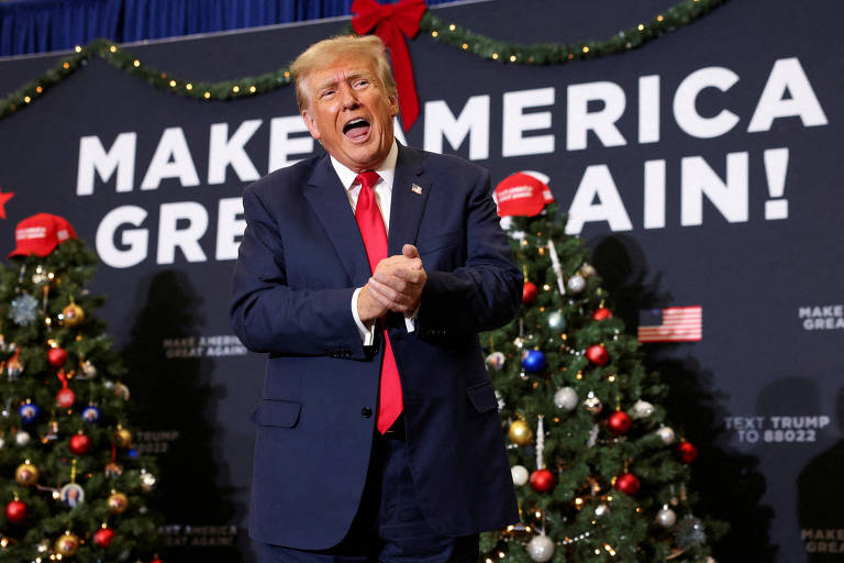 Homem idoso branco, de cabelos loiros, vestindo terno azul e gravata vermelha, aparece em palco com os dizeres "Make America Great Again"ao fundo