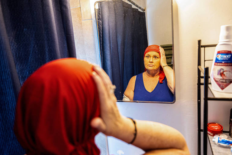 Fotografia colorida mostra uma mulher branca se olhando no espelho; ela olha se reflexo enquanto ajusta um lenço vermelho na cabeça