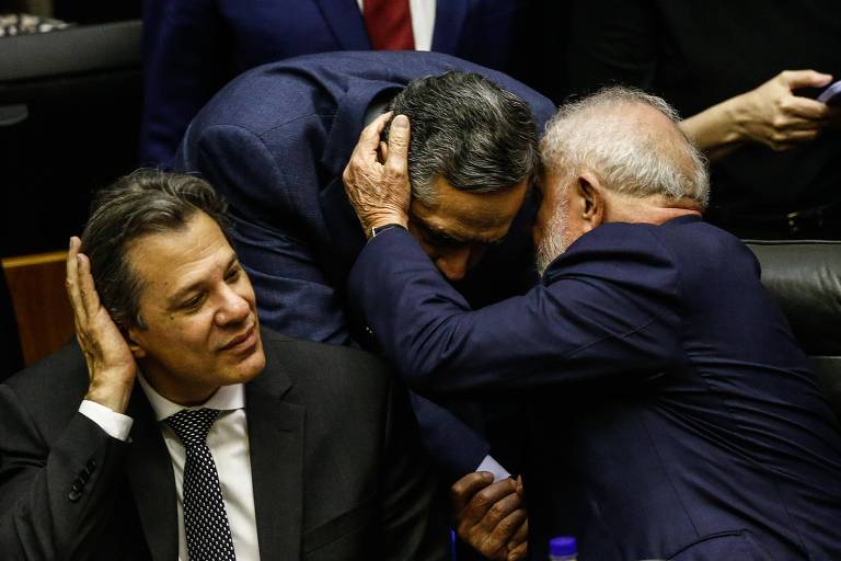 Imagem mostra Lula falando ao ouvido de Barroso. Ao lado, Haddad mexe no cabelo com uma das mãos.