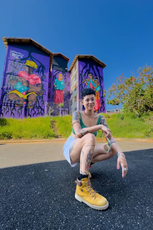 Mulher jovem, com corpo tatuado, posa em frente aos prédios grafitados; ela usa miniblusa, shorts e botas amarelas