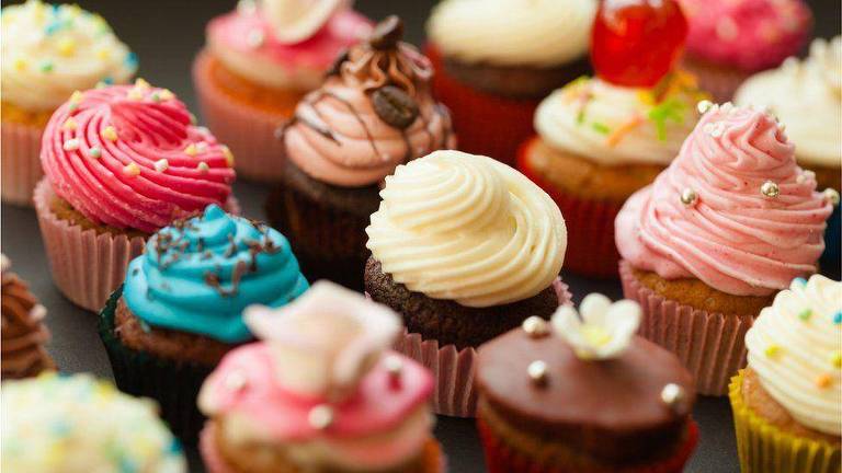 Foto mostra vários cupcakes (pequenos bolinhos) com coberturas coloridas (em sua maioria, rosa, azul e amarelo) e confeitos